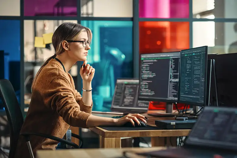 Eine Programmiererin begutachtet Code auf ihrem Monitor vor bunten Glaswänden