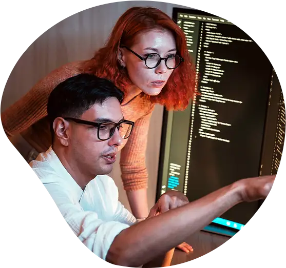 Zwei Softwareentwickler arbeiten an zwei Monitoren zusammen und überprüfen Code in einem schwach beleuchteten Büro; einer zeigt auf den Bildschirm, während der andere aufmerksam zusieht.