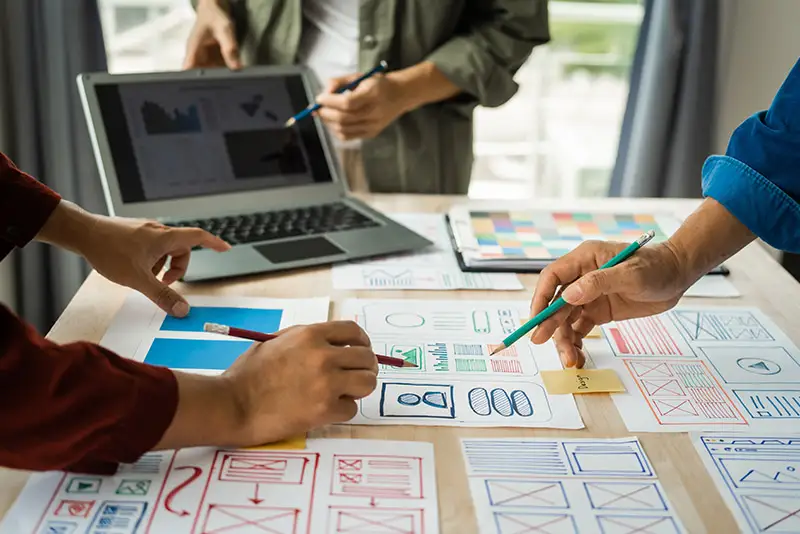 Mitglieder eines Projektteams diskutieren über einem Tisch mit Mockups und Laptop