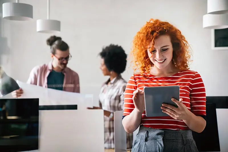 Eine junge Frau betrachtet zufrieden ihr Tablet, im Hintergrund ein Büro