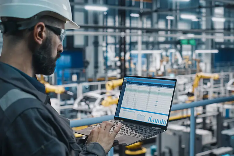 Ein Industriearbeiter mit Schutzhelm betrachtet die Prozesse an einem Laptop in einer Produktionshalle